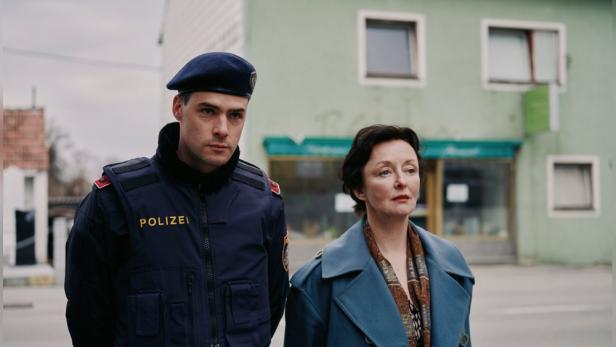 Ein Polizist in Uniform steht neben einer Frau mit blauem Mantel vor einem Gebäude mit grüner Fassade.