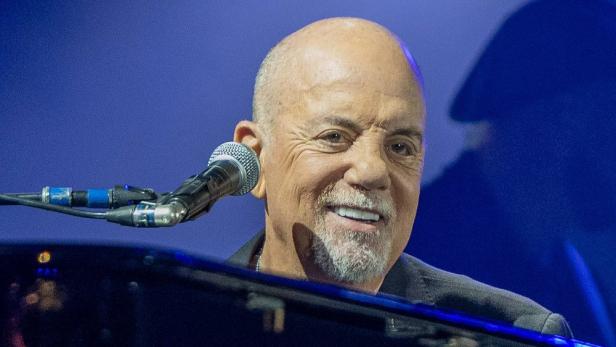 Billy Joel lächelnd am Klavier neben Mikrofon vor blauem Hintergrund.