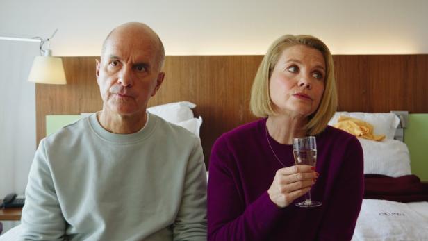 Ein älteres Paar sitzt nebeneinander auf einem Bett, beide schauen ernst, die Frau hält ein Glas mit Getränk.