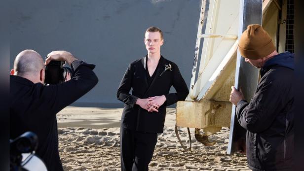 Eine Person in schwarzer Kleidung posiert am Strand neben einer Metallstruktur, während zwei Männer das Fotoshooting unterstützen.
