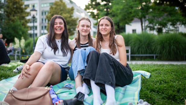 Drei junge Frauen sitzen lachend auf einer Picknickdecke im Park, umgeben von Grün und Gebäuden im Hintergrund.