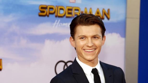 Tom Holland lächelt vor einem "Spider-Man"-Hintergrund in einem dunklen Anzug.