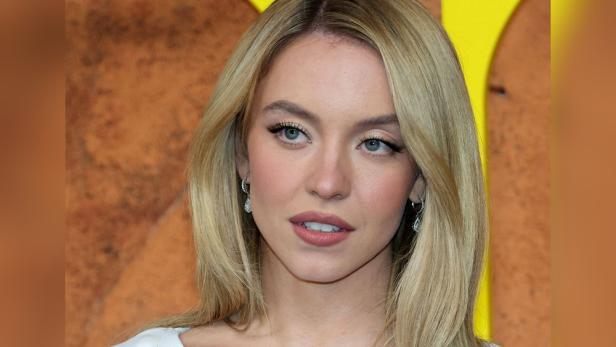Sydney Sweeney mit blonden, schulterlangen Haaren und Ohrringen vor gemustertem Hintergrund.
