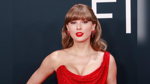 Taylor Swift trägt ein rotes Kleid und roten Lippenstift vor einem dunklen Hintergrund.