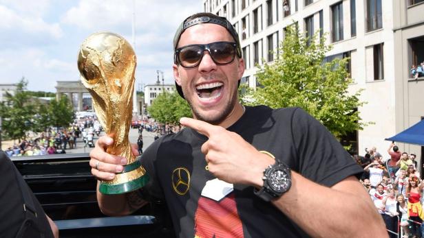 Ein jubelnder Lukas Podolski mit Sonnenbrille hält den WM-Pokal in der Hand und zeigt darauf.