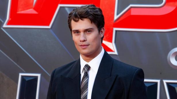 Nicholas Galitzine trägt einen schwarzen Anzug mit weißem Hemd und gestreifter Krawatte vor einem roten Hintergrund.