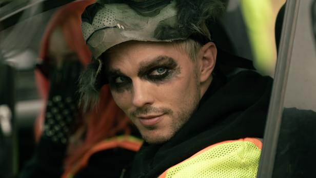 Ein junger Mann mit dunklem Augen-Make-up und einer Wolfsmaske auf dem Kopf sitzt in einer Warnweste in einem Auto.