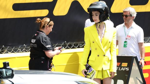Lily Collins trägt einen gelben Hosenanzug und einen Rennhelm an einer Formel-1-Strecke, während eine Cadillac-Mitarbeiterin daneben steht.