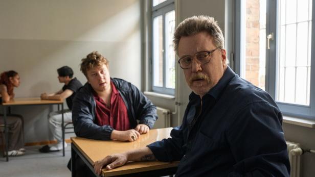 Ein Mann mit Schnurrbart und Brille sitzt in einem Raum mit vergitterten Fenstern, während im Hintergrund zwei weitere Personen an einem Tisch sitzen.