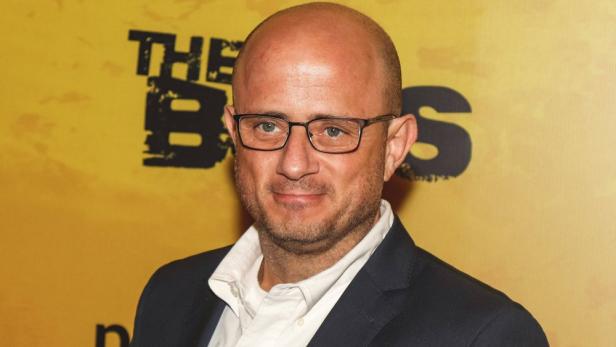 Ein Mann mit Brille und Glatze trägt ein dunkles Sakko und steht vor einem gelben Hintergrund mit schwarzer Schrift.