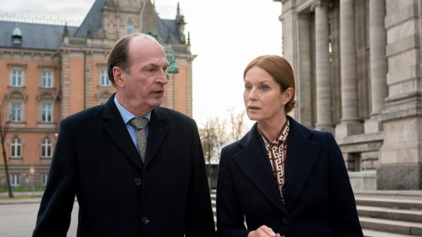 Ein Mann und eine Frau in dunklen Mänteln unterhalten sich vor einem historischen Gebäude.