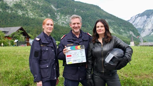Zwei Polizisten und eine Frau mit Motorradhelm stehen vor einer Bergkulisse und halten eine Filmklappe für die Produktion „Letzter Tropfen“.