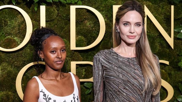 Angelina Jolie posiert mit ihrer Tochter Zahara Jolie-Pitt vor einer mit Moos bewachsenen Wand mit goldenem Schriftzug.
