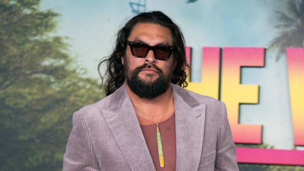 Jason Momoa trägt eine Sonnenbrille, ein mauvefarbenes Cord-Sakko und eine lange Halskette vor einem bunten Hintergrund.