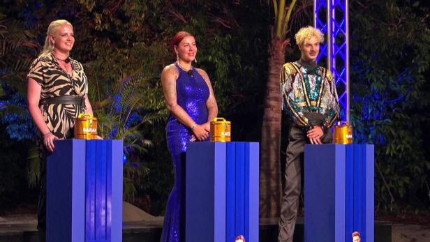 Drei Personen stehen nebeneinander an blauen Podesten mit goldenen Behältern, im Hintergrund sind Pflanzen und bunte Beleuchtung.