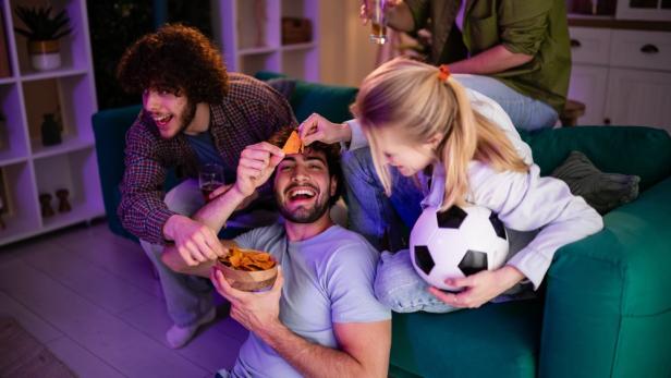 Eine Gruppe junger Freunde sitzt auf einem Sofa, isst Chips und schaut gemeinsam Fußball.
