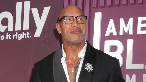 Dwayne Johnson trägt eine Brille und einen schwarzen Anzug mit weißem Hemd vor einem violetten Hintergrund.