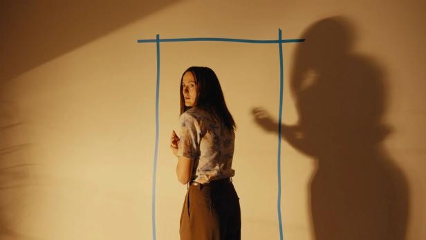 Eine Frau blickt ängstlich über die Schulter, während ihr Schatten neben einem blauen, auf die Wand geklebten Rechteck eine bedrohliche Geste macht.