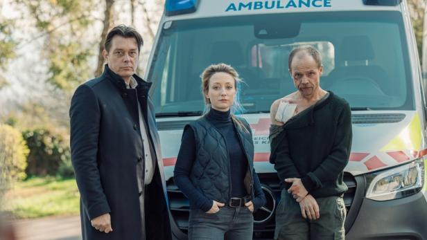 Drei Personen stehen vor einem Krankenwagen, eine davon hat eine sichtbare Verletzung an der Schulter.