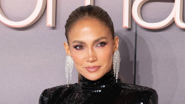 Jennifer Lopez trägt ein schwarzes Paillettenoberteil und auffällige, lange Strass-Ohrringe vor einem grauen Hintergrund.