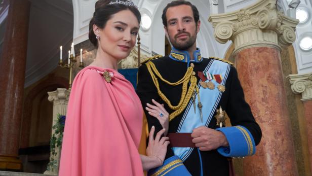 Eine Frau in einem rosa Kleid und ein Mann in einer prunkvollen Militäruniform stehen gemeinsam in einem prachtvollen Saal.