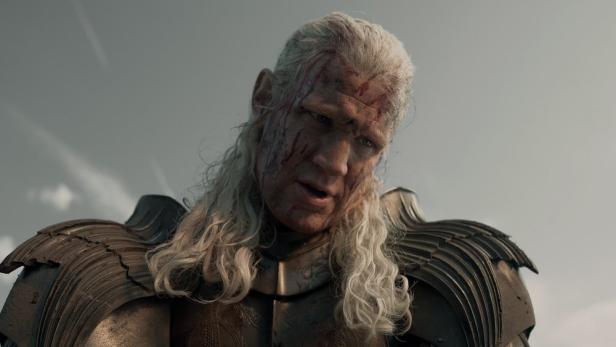 Matt Smith als Daemon Targaryen mit blutverschmiertem Gesicht und langem, hellem Haar in einer Rüstung vor einem grauen Himmel.