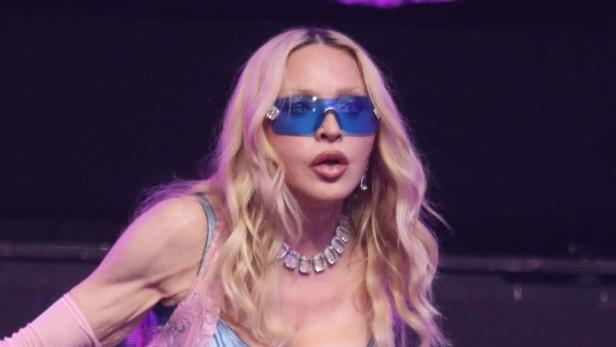 Madonna trägt eine blaue Sonnenbrille und eine auffällige Halskette vor einem dunklen Hintergrund.