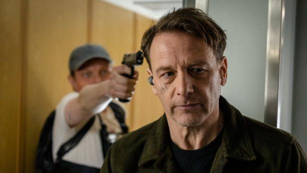 Ein Mann mit ernstem Blick steht im Vordergrund, während im Hintergrund eine Person mit einer Pistole auf ihn zielt.