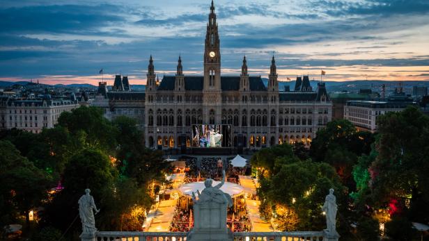 Das Wiener Rathaus bei Abenddämmerung mit einer großen Leinwand und beleuchteten Ständen auf dem Rathausplatz.