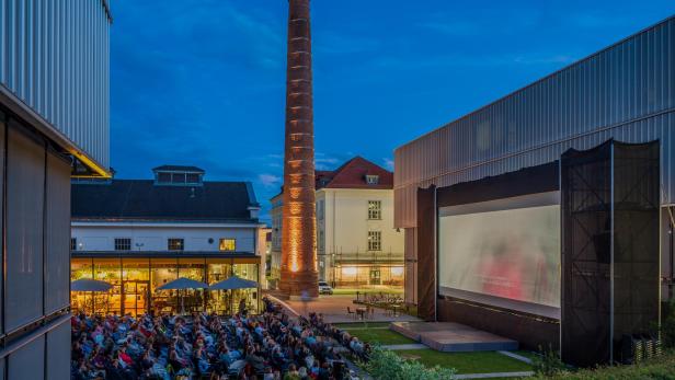 Ein Publikum schaut abends auf einer Freilichtbühne vor einem beleuchteten historischen Backsteinschornstein einen Film.