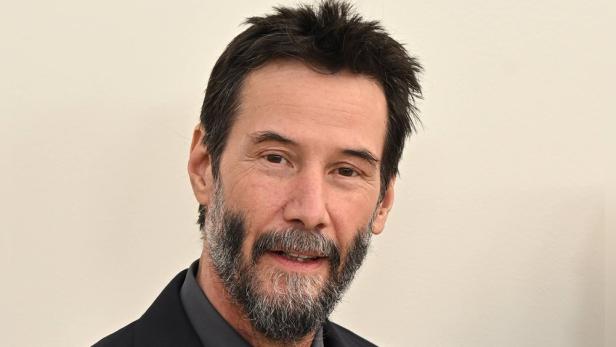 Keanu Reeves mit dunklem Haar und Bart vor einem hellen Hintergrund.