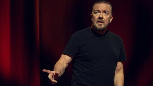 Ricky Gervais steht in einem schwarzen T-Shirt vor einem roten Vorhang und zeigt mit dem Finger zur Seite.