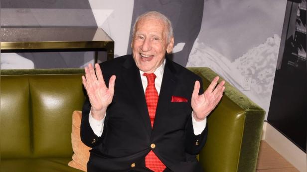 Mel Brooks sitzt lächelnd mit erhobenen Händen auf einem grünen Sofa und trägt einen schwarzen Anzug mit roter Krawatte.
