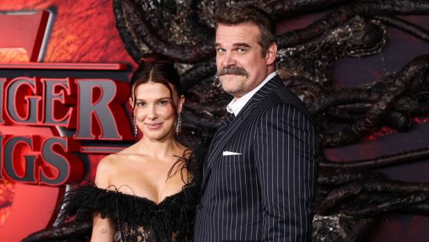 Millie Bobby Brown und David Harbour posieren vor einem „Stranger Things“-Hintergrund.