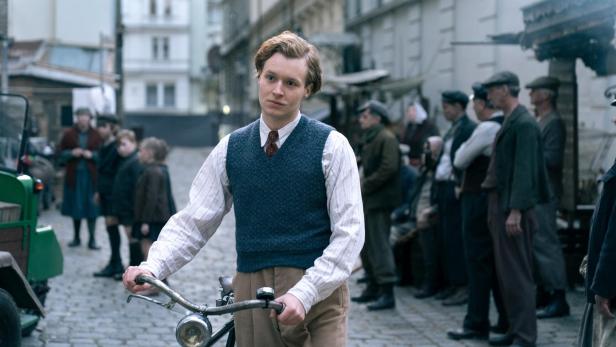Ein junger Mann in historischer Kleidung schiebt ein Fahrrad durch eine belebte Gasse mit Kopfsteinpflaster.