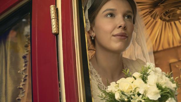 Millie Bobby Brown schaut als Enola Holmes in einem weißen Brautkleid mit Schleier und Blumenstrauß aus dem Fenster einer Kutsche.