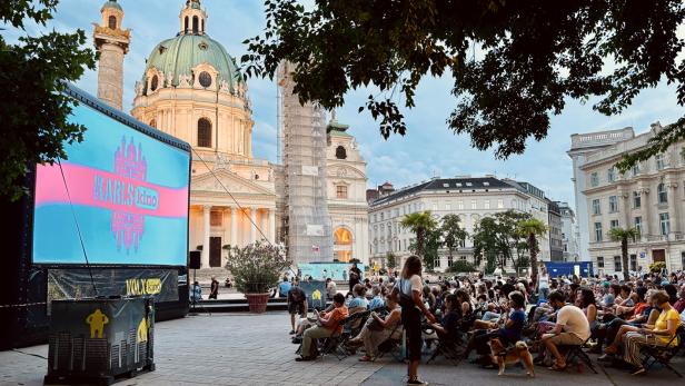 Zuschauer sitzen vor einer großen Leinwand des Volxkino auf dem Wiener Karlsplatz mit der Karlskirche im Hintergrund.