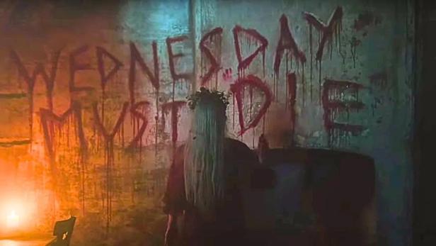 Eine Frau mit langem blonden Haar vor einer Wand, auf der in blutroter Schrift „WEDNESDAY MUST DIE“ steht.