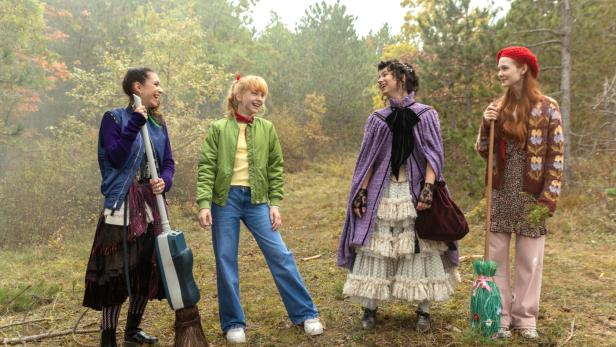 Vier junge Frauen in unterschiedlichen Outfits stehen lachend in einem herbstlichen Wald, wobei zwei von ihnen Besen halten.
