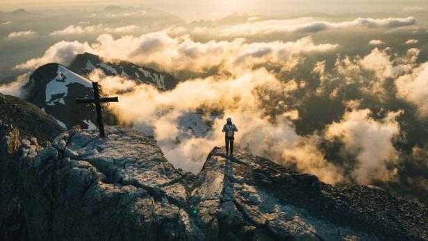 Eine Person steht auf einem felsigen Berggipfel neben einem Gipfelkreuz über einem Meer aus Wolken im warmen Licht der untergehenden Sonne.