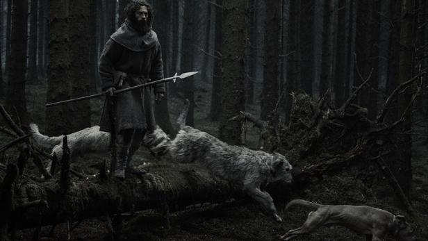 Ein bärtiger Mann in mittelalterlicher Kleidung mit einem Speer steht in einem dunklen Wald, während zwei Hunde über einen umgestürzten Baum springen.