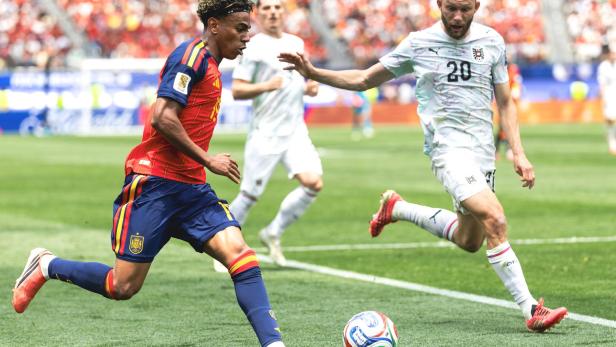 Ein spanischer Spieler im roten Trikot führt den Ball gegen einen österreichischen Gegenspieler mit der Nummer 20 auf dem Rasen.