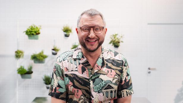 Ein lächelnder Mann mit Brille trägt ein buntes Hemd mit Comic-Motiven vor einer Wand mit hängenden Pflanzen.