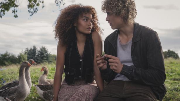 Eine junge Frau mit lockigem Haar und ein junger Mann mit blonden Locken sitzen sich auf einer Wiese mit Gänsen gegenüber und schauen einander an.