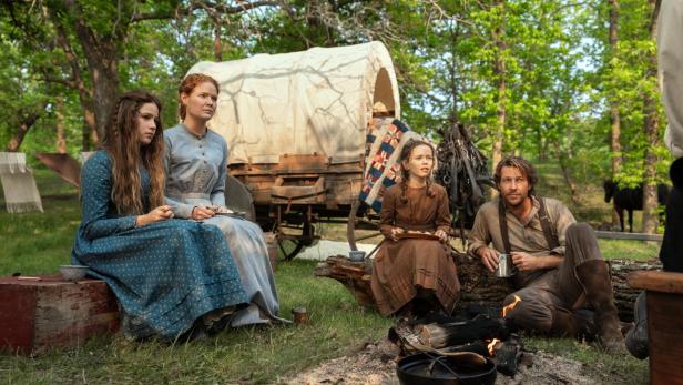 Eine Familie in historischer Kleidung sitzt vor einem Lagerfeuer neben einem Planwagen in einem Wald.