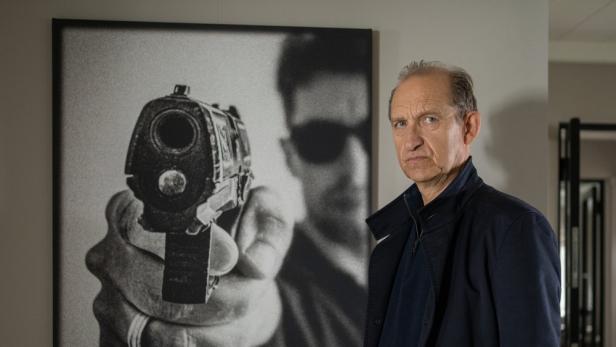 Ein älterer Mann mit ernstem Blick steht vor einem schwarz-weißen Wandbild, das eine Person mit einer auf den Betrachter gerichteten Pistole zeigt.