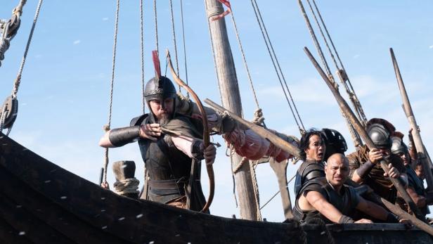 Ein Krieger mit Helm zielt auf einem antiken Holzschiff mit einem Bogen, während weitere Kämpfer das Boot steuern.