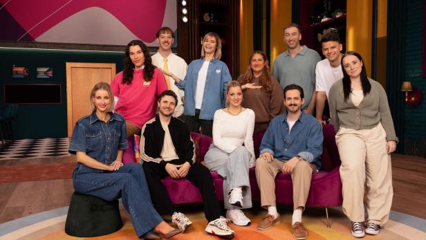 Eine Gruppe von elf jungen Erwachsenen posiert gemeinsam in einem farbenfrohen Fernsehstudio vor einem violetten Sofa.