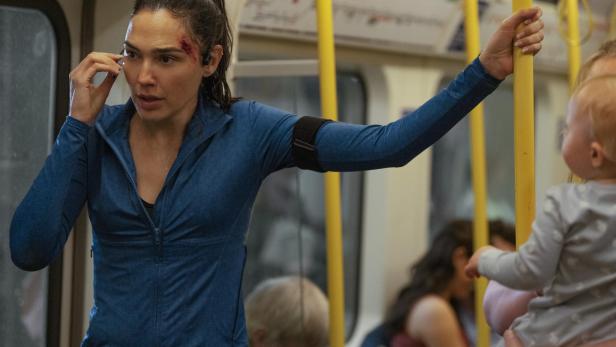 Gal Gadot mit einer Kopfverletzung telefoniert in einer U-Bahn, während sie sich an einer gelben Stange festhält.