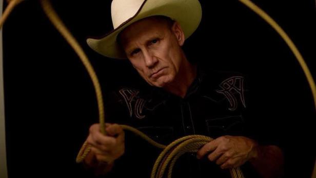 Ein Mann mit Cowboyhut und schwarzem Hemd hält vor einem dunklen Hintergrund ein Lasso in den Händen.